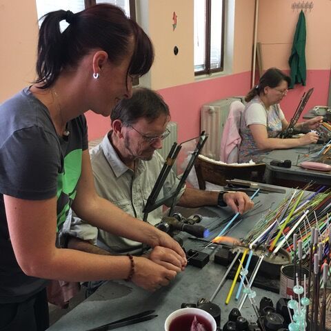 At a lampwork bead workshop, a demonstrator leans in to guide Matthew as he shapes glass in the torch flame, surrounded by colourful glass rods, while another guest makes beads in the background.